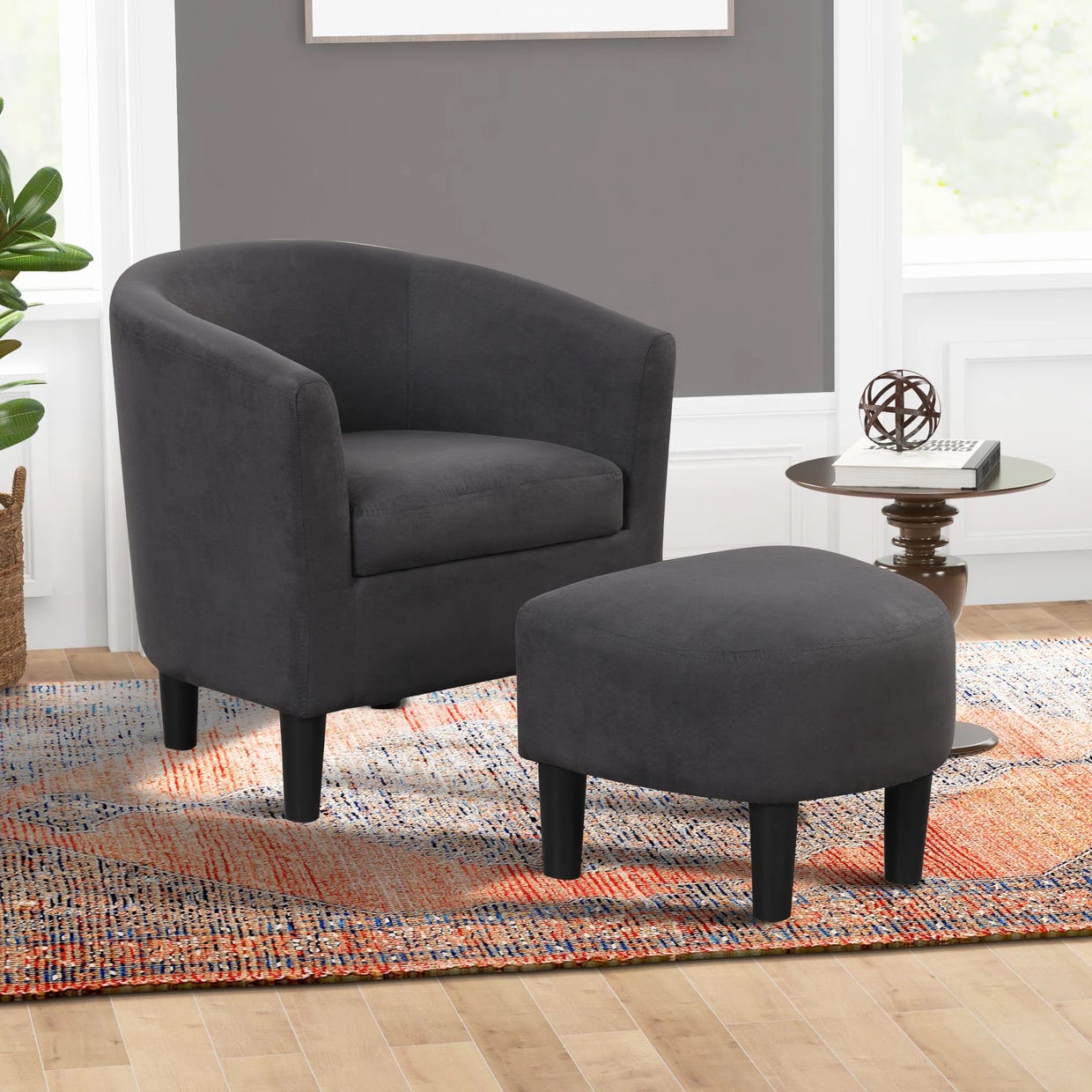 A dark grey upholstered armchair with a matching ottoman, set on a multicolored area rug beside a round side table with a decorative object.