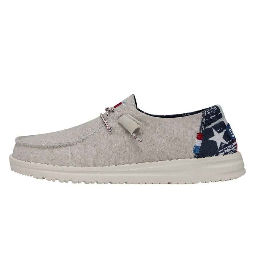 Hey Dude Wendy Texas Canvas is a casual shoe with a light beige fabric upper, featuring star and flag patterns on the heel, lace-up closure, and a cushioned white sole for comfort.