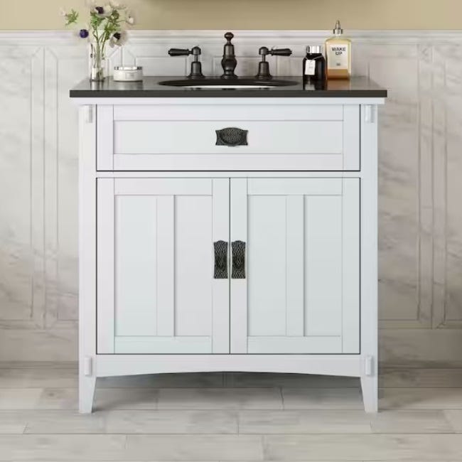 White bathroom vanity with a black countertop, equipped with a sink and faucet; beneath, there's a cabinet with doors and a drawer.