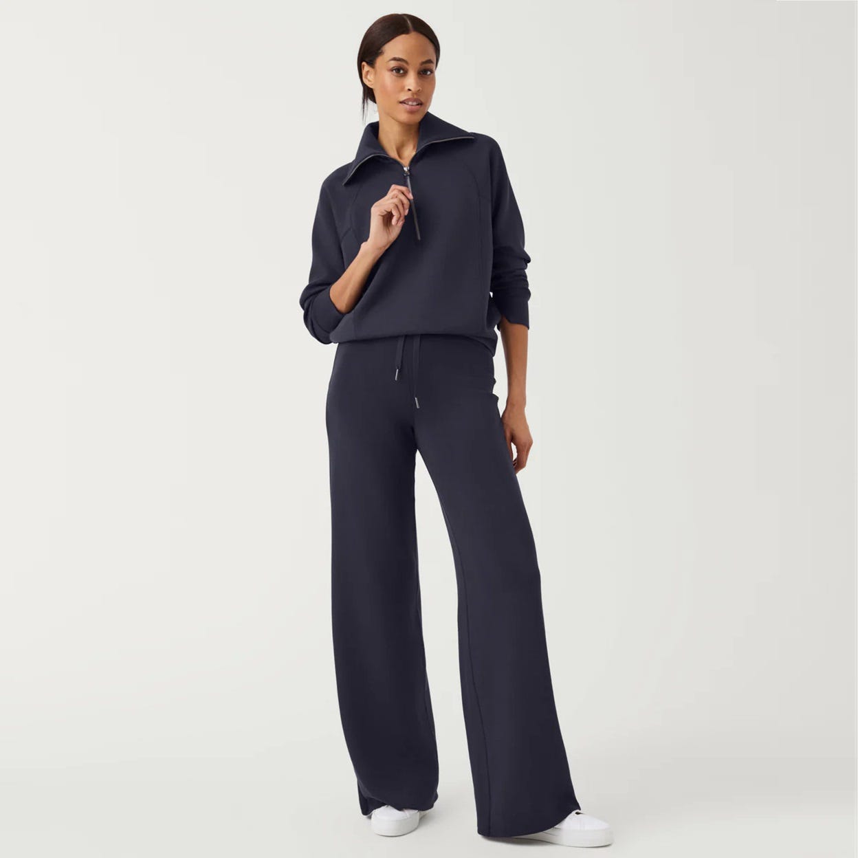 A woman models a navy blue tracksuit with a half-zip sweatshirt and wide-leg pants, paired with white sneakers.