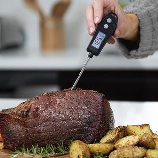 A cooked roast beef is being checked with a digital meat thermometer; roasted potatoes accompany the dish.