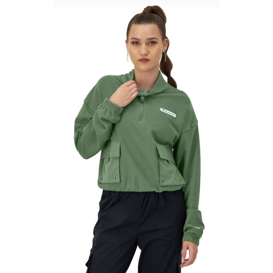 A woman is wearing a green half-zip pullover with front pockets and black pants.