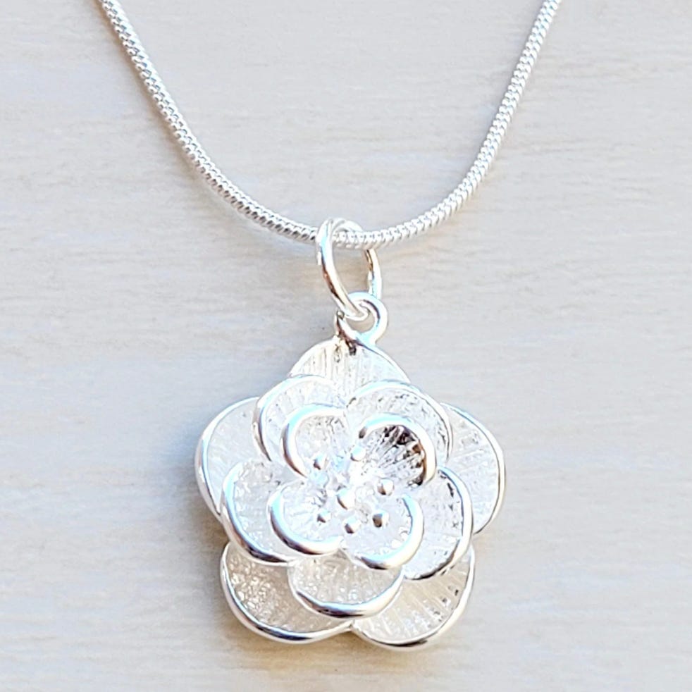 A silver necklace with a layered flower pendant hangs against a light wooden background. The flower design features intricate cut-out patterns.