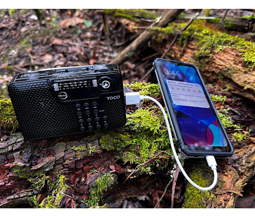 A portable radio and a smartphone connected by a charging cable outdoors on a log.