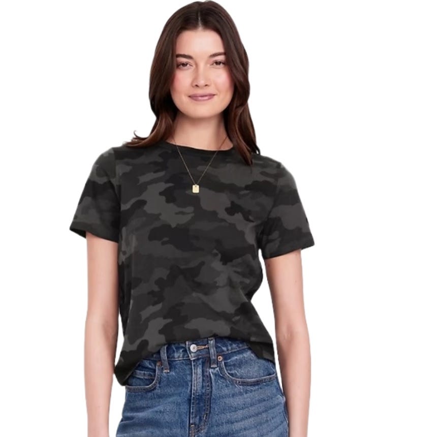 A person wearing a dark camouflage t-shirt with short sleeves and blue jeans, accessorized with a small pendant necklace.