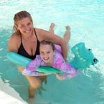 A woman and a child are floating in a pool using a dinosaur-shaped inflatable and a printed swim ring, respectively.
