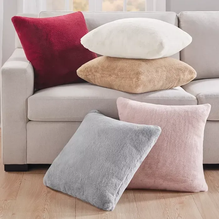 The photo shows a collection of fluffy throw pillows in varying colors including red, white, tan, gray, and pink on a beige sofa.