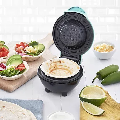 A teal and black Dash Mini Waffle Bowl Maker is open, displaying its waffle bowl-shaped cooking plates; ingredients for taco bowls are spread around it.