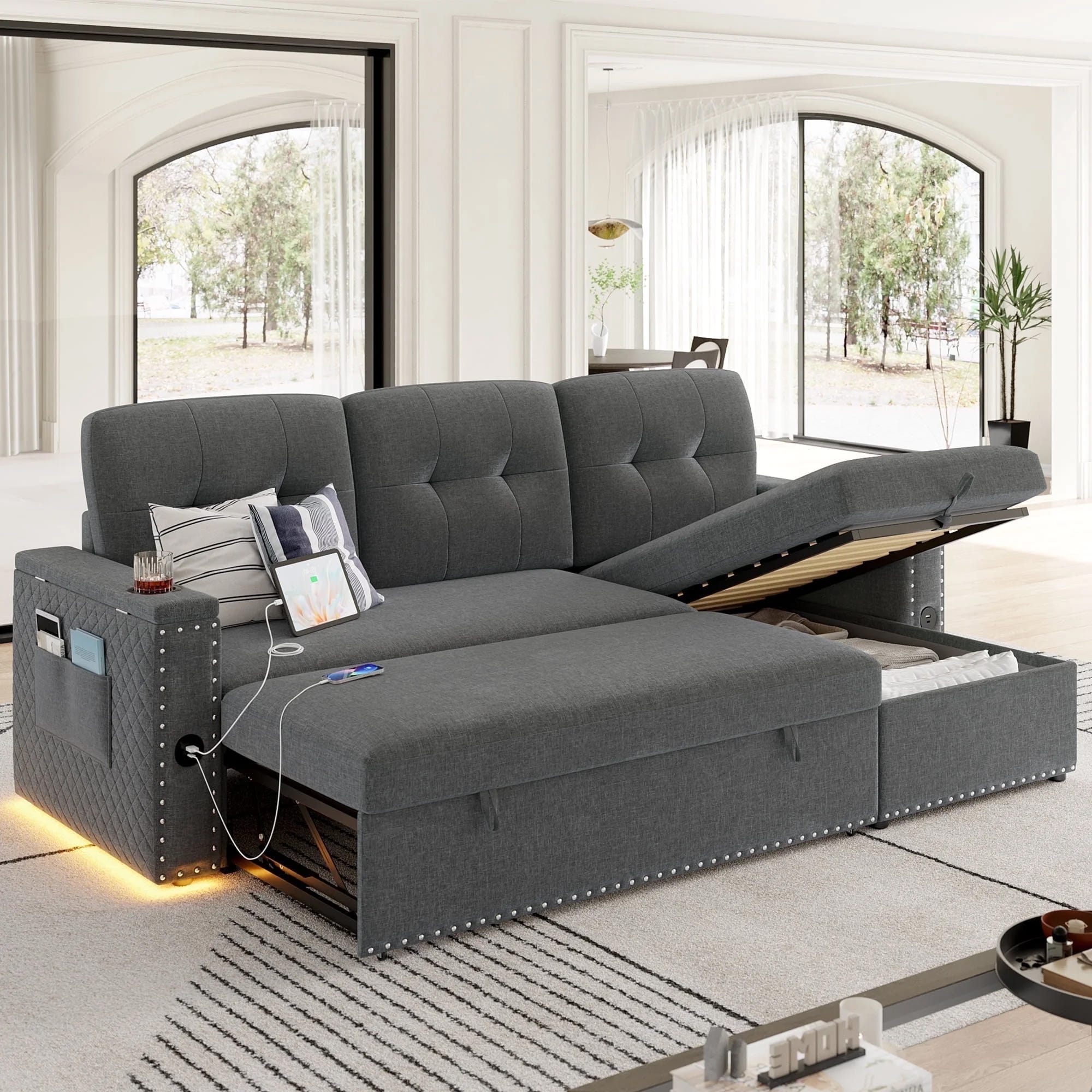A gray sectional sofa with a pull-out bed and storage compartment. It features stud detailing, built-in lights, and a charging station with pockets for devices and magazines.