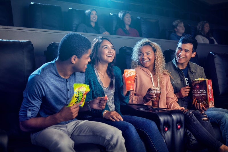 Four friends are enjoying a movie in a theater, holding soft drinks and popcorn.