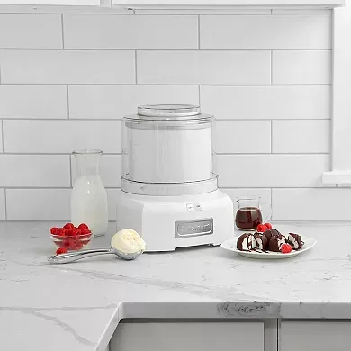 A Cuisinart Ice Cream Maker sits on a kitchen countertop accompanied by ingredients like milk, raspberries, and a bowl of ice cream with toppings.