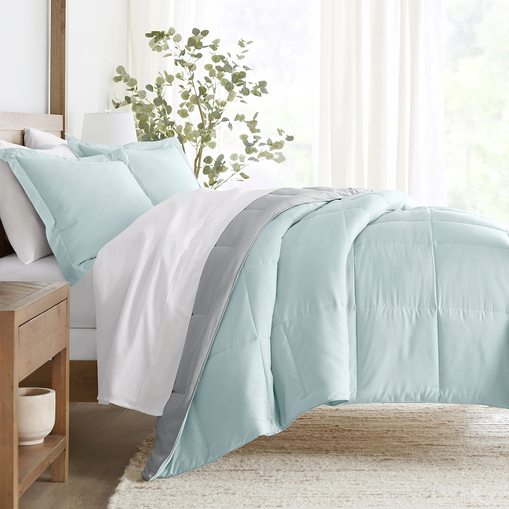 3pc Queen Reversible Comforter Set features a light blue comforter with a contrasting gray reversible side and matching pillow shams. Set is styled on a bed with white sheets in a bright, airy room with a wooden side table and green plant.