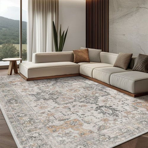 A large 8'x10' rug featuring a detailed, intricate vintage pattern in muted tones of gray, beige, and subtle orange. It lies on a wooden floor in a modern living room with a sectional sofa, a side table, and greenery in the background.