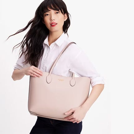 Woman holding a large beige tote bag and wearing a white button-up shirt with dark jeans.