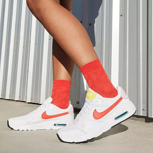 A pair of white Nike Air Max SC sneakers with contrasting red Swoosh logos and orange Nike branding on the tongue, featuring a visible Air Max cushioning unit in the heel.