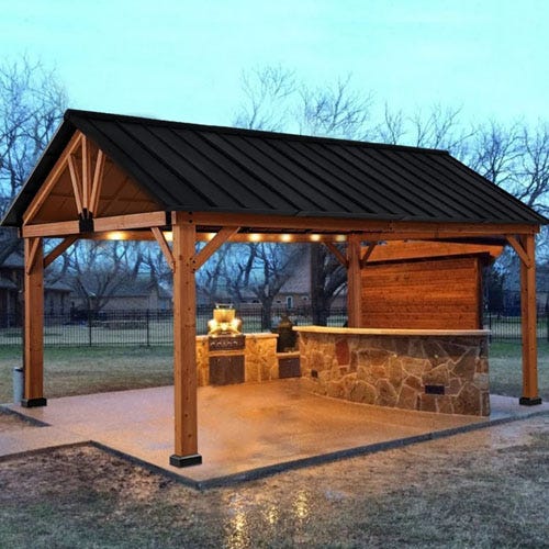 An outdoor pavilion with a stone bar and built-in grill under a metal roof.