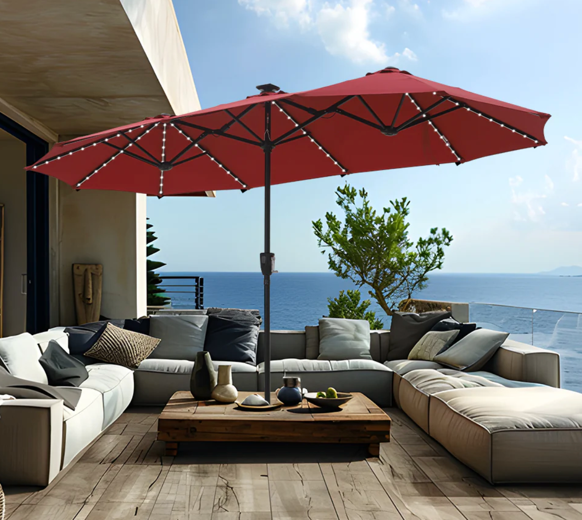 The photo shows a red patio umbrella, an L-shaped outdoor sectional sofa, and a wooden coffee table on a deck overlooking the sea.