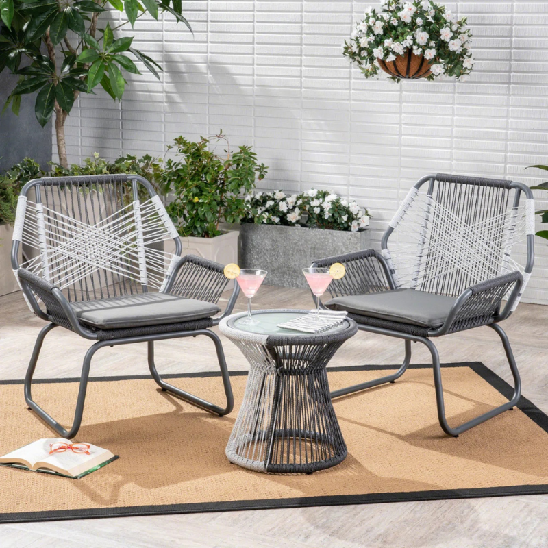 Outdoor patio set with two woven chairs and a small round table. The setting includes a beige rug, greenery, flowers, an open book, and two pink cocktails with lemon slices.
