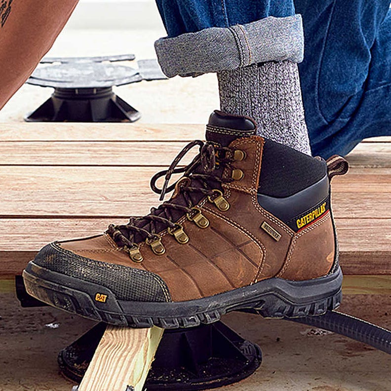 A pair of brown work boots with a person wearing gray socks and blue jeans.