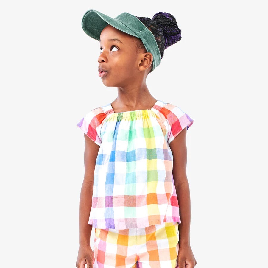 A young girl wearing a colorful checked outfit and a green cap.