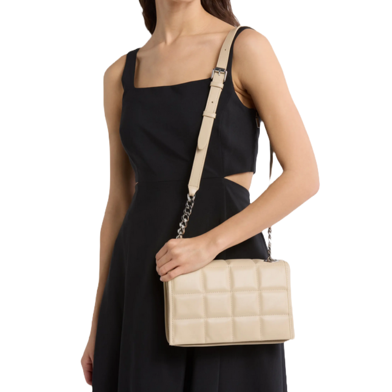 A woman in a black sleeveless dress holds a beige quilted handbag with an adjustable strap and chain detail.