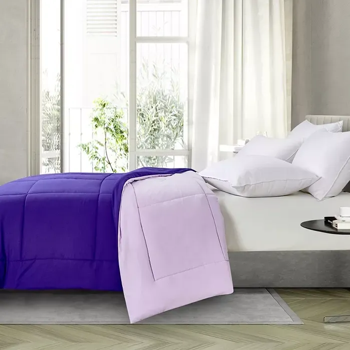 A bed with a white sheet and pillowcases, covered by a purple comforter and a folded lighter purple blanket at the foot.