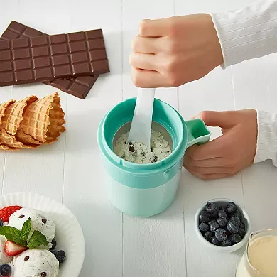 A person is mixing ingredients inside a compact, turquoise ice cream maker with a white mixing spoon, with waffle cones, a bar of chocolate, and small bowls of strawberries and blueberries nearby.