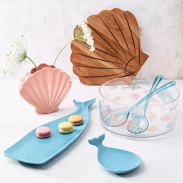 Various sea-themed serving items: a pink shell-shaped vase, a leaf-shaped wooden tray, a blue fish platter with macarons, and a clear bowl with fish patterns and blue tongs.