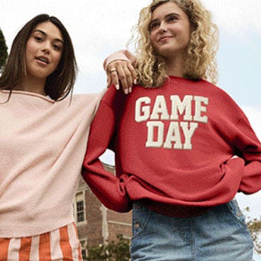 Two people are wearing casual outfits: one in a pink sweater with striped shorts, and the other in a red sweatshirt with \“GAME DAY\“ text paired with a denim skirt.