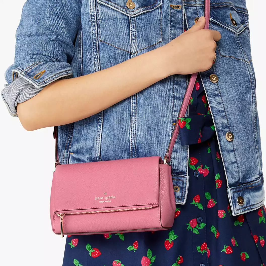 A pink crossbody purse with a front zipper, worn with a denim jacket and a patterned dress.