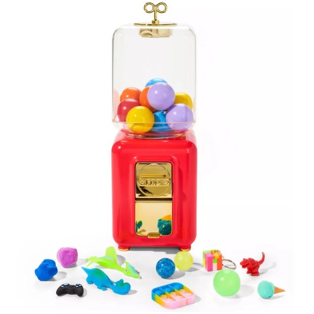 A vintage-style red and gold vending machine with colorful plastic balls dispenses small toys like dinosaurs, a game controller, a ball, and a tiny gift box scattered in front.