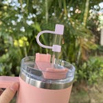 A silicone straw topper in the shape of a heart with a slot for a straw is attached to a clear straw in a pink insulated tumbler.