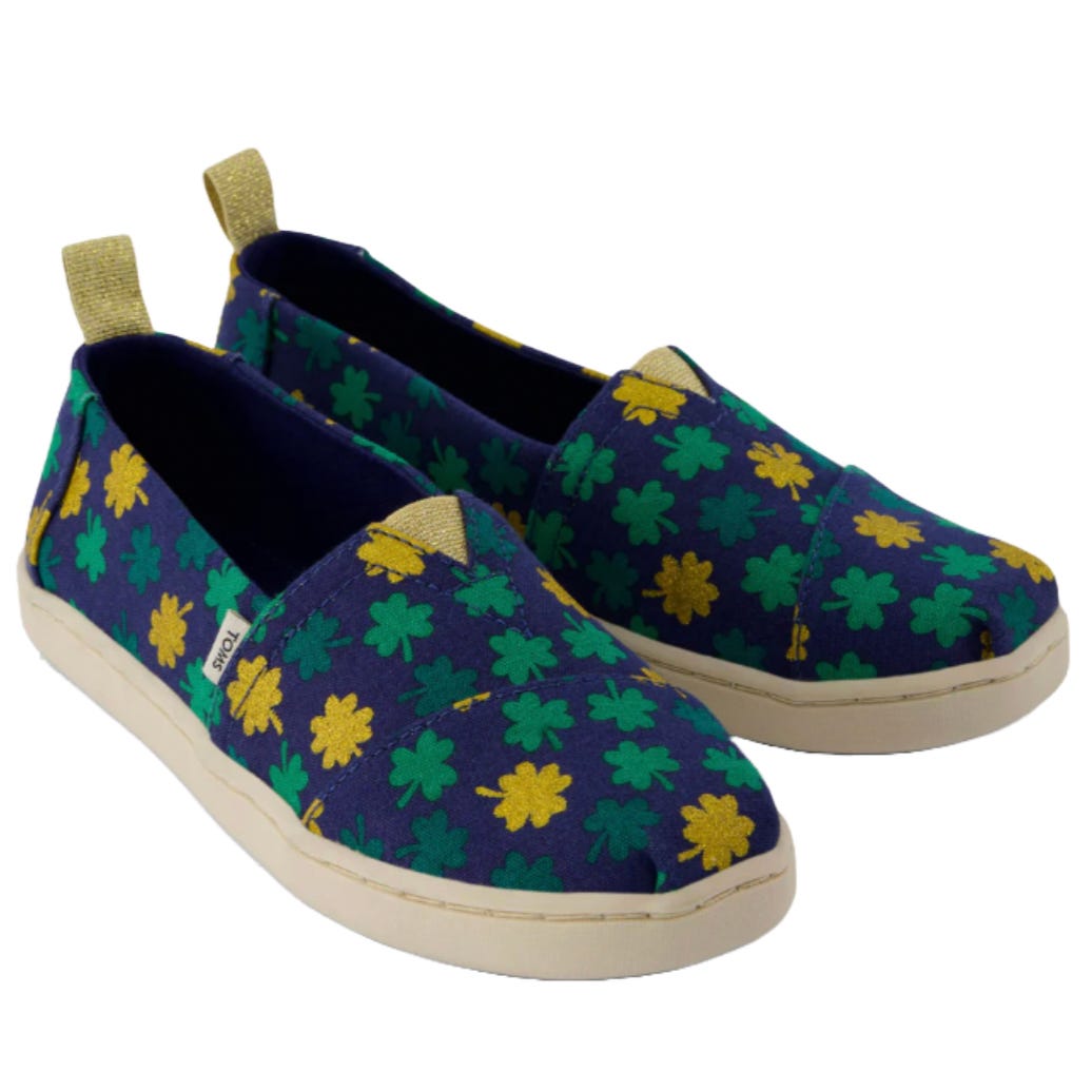 Kids' slip-on shoes featuring a navy blue fabric adorned with green and yellow clover patterns, complemented by a beige pull tab and sole.
