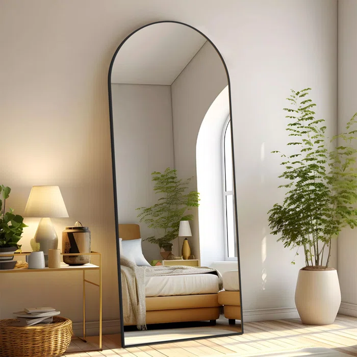 A modern bedroom features a large, arched floor mirror, a cozy bed with a beige headboard, a side table with a lamp and decor, and a potted plant, all set against a neutral color palette with natural light streaming in.