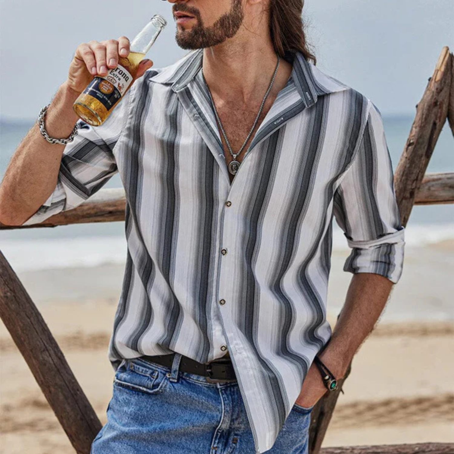 A man is wearing a Coofandy shirt featuring vertical black, white, and gray stripes with a button-up front and long sleeves rolled up to the elbows, paired with blue jeans. He has a necklace and is holding a bottle near the beach.