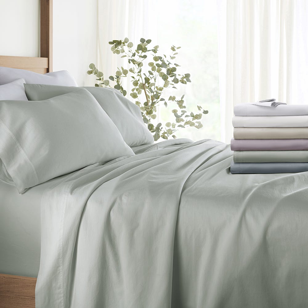 Bed with light green sheets and a stack of folded sheets in various colors beside it.