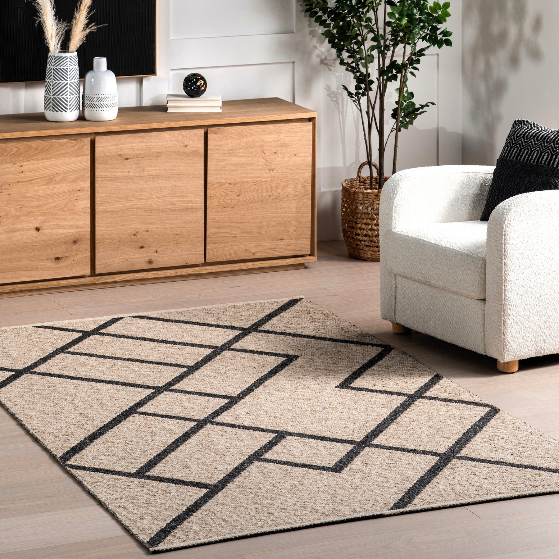 A cozy living room setup featuring a black and cream geometric-patterned rug, a white armchair with a black cushion, a wooden sideboard, decorative vases, and a potted plant.