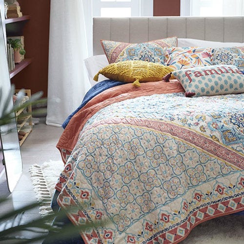 A patterned quilt, matching pillows, and a yellow cushion on a bed with a beige headboard.