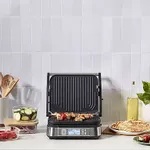 A Cuisinart Griddler is shown open, with angled grill plates, digital controls, and various foods being prepped in the background.