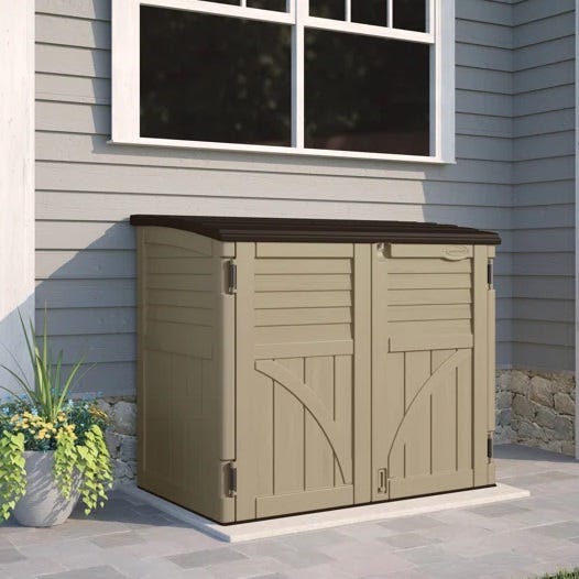 A beige outdoor storage shed with a brown roof, situated beside a house with a window.