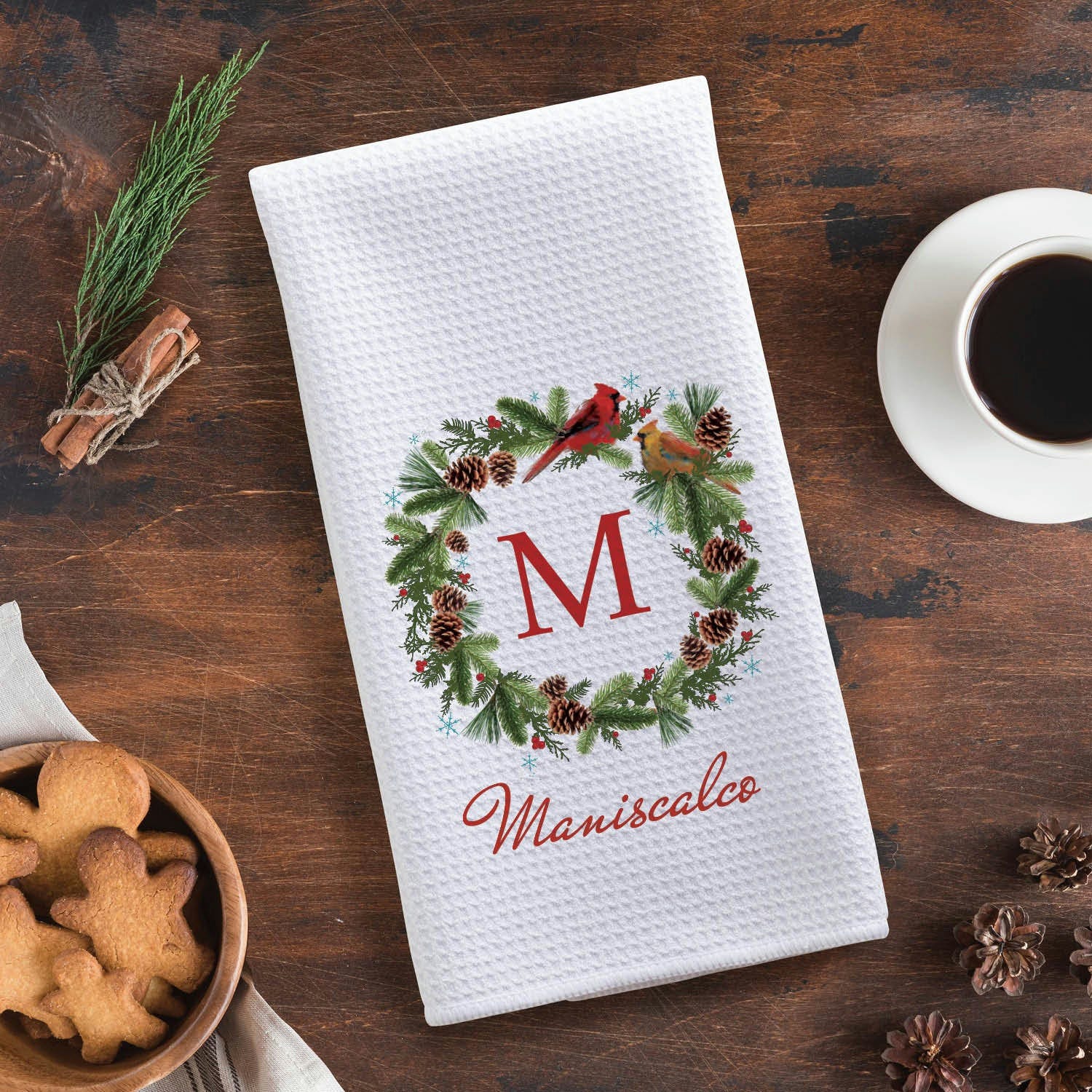 A white kitchen towel featuring a festive wreath design with pinecones, greenery, and two birds, centered with the letter \“M\“ and the word \“Maniscalco\“ below. It is placed on a wooden surface with a cup of coffee, cookies, and cinnamon sticks nearby.