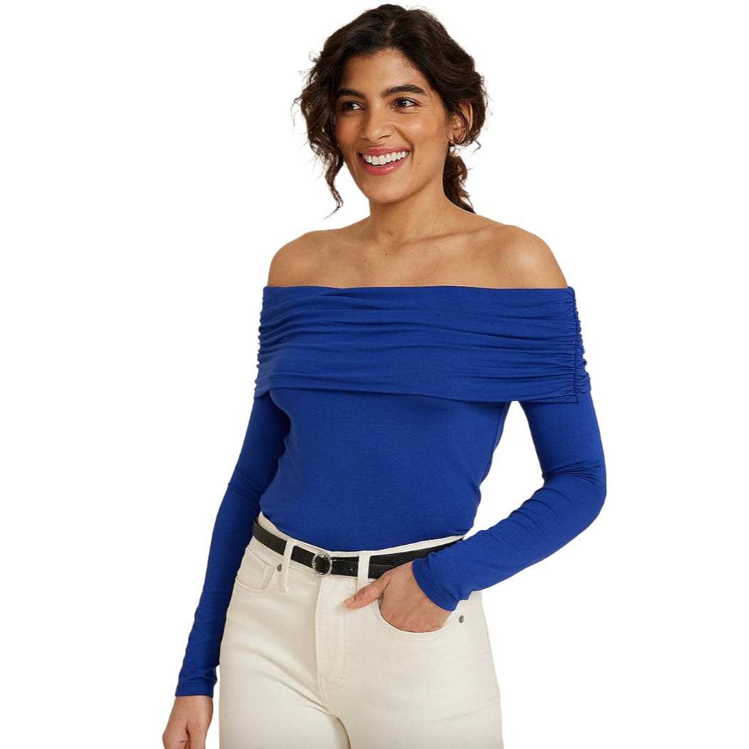 A woman is wearing an off-the-shoulder blue top with long sleeves paired with white pants and a black belt. She's smiling.