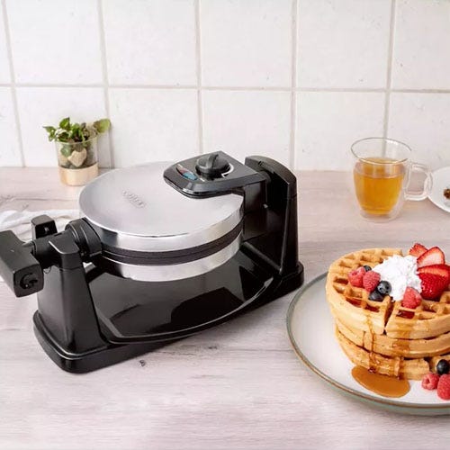 A waffle maker with a finished waffle topped with cream and berries on a plate, next to a cup of tea.