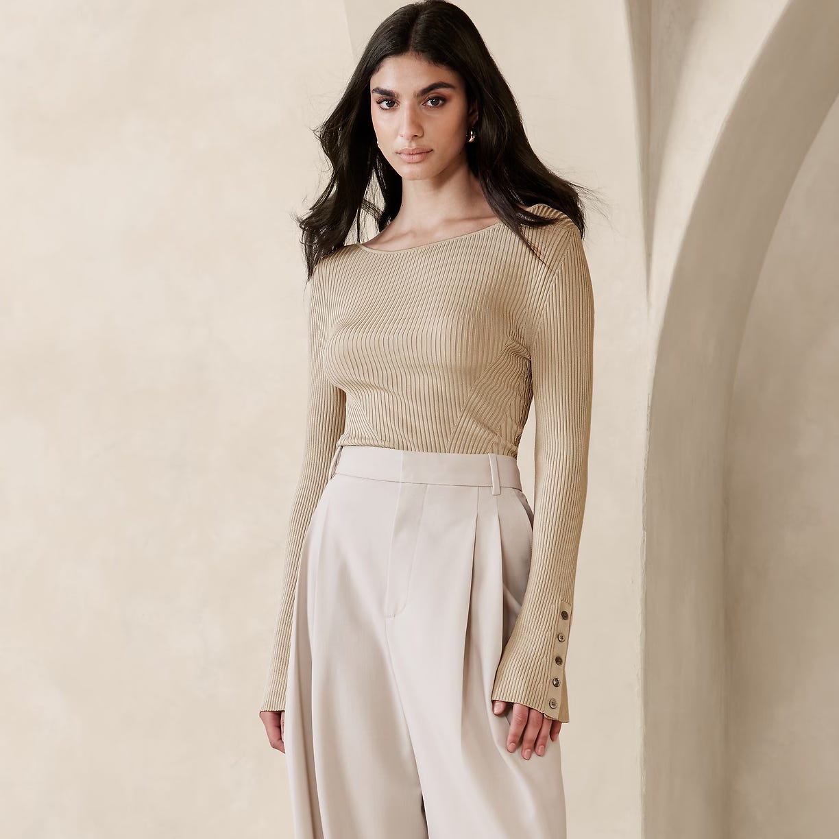 A woman is wearing a beige ribbed sweater and high-waisted cream trousers.