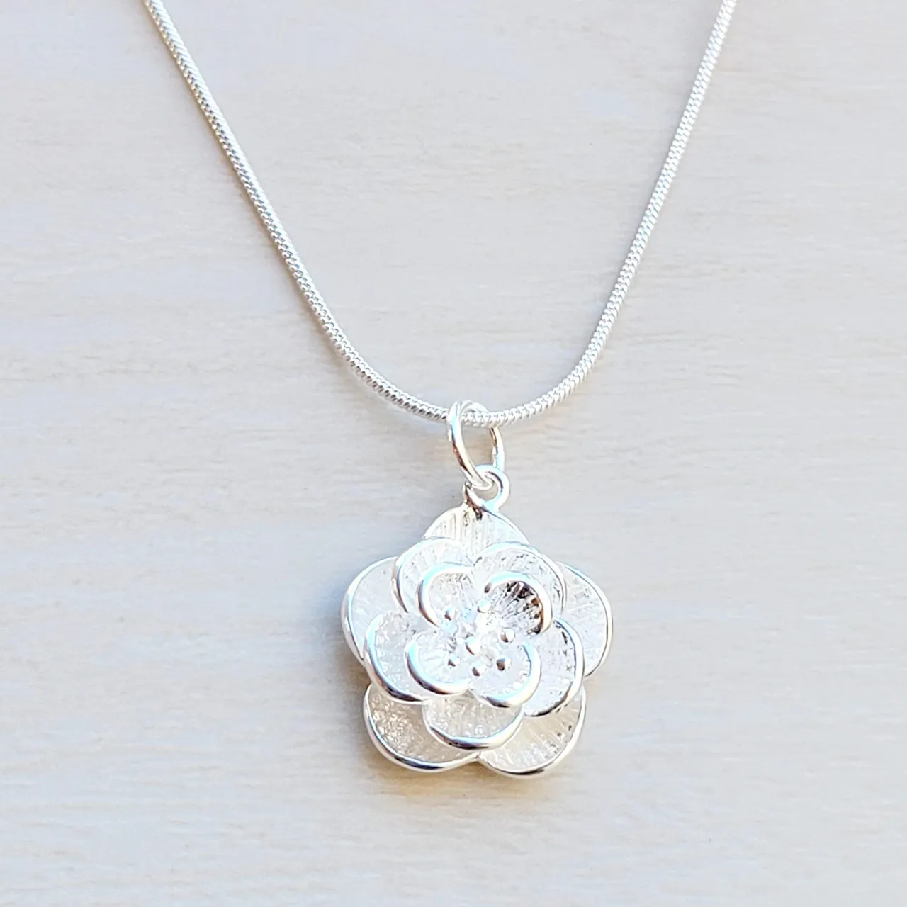 A silver necklace features a pendant designed as a layered flower with textured petals, attached to a sleek chain.