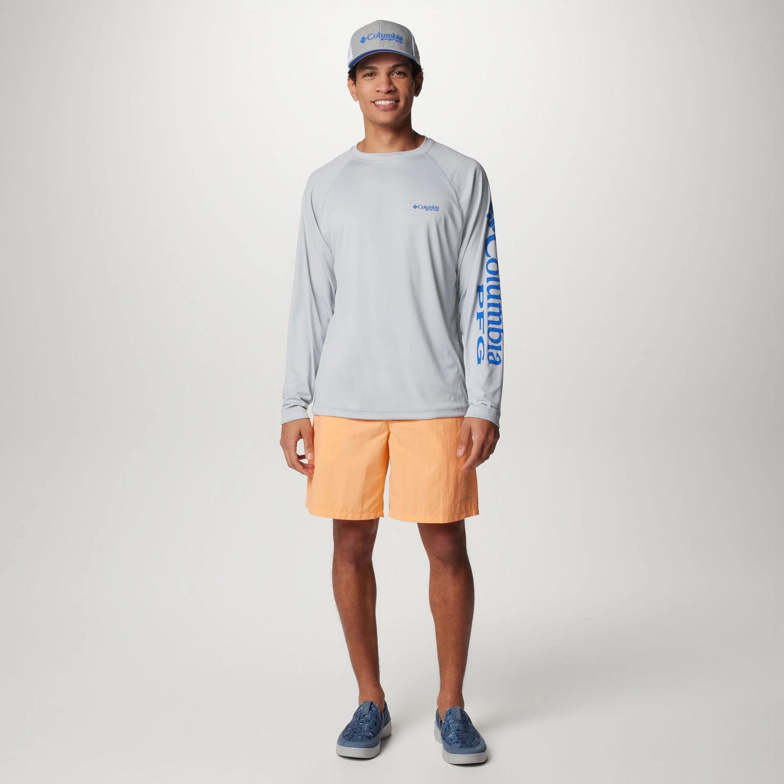 A man models a grey long-sleeve shirt, orange shorts, a white cap, and blue sneakers.
