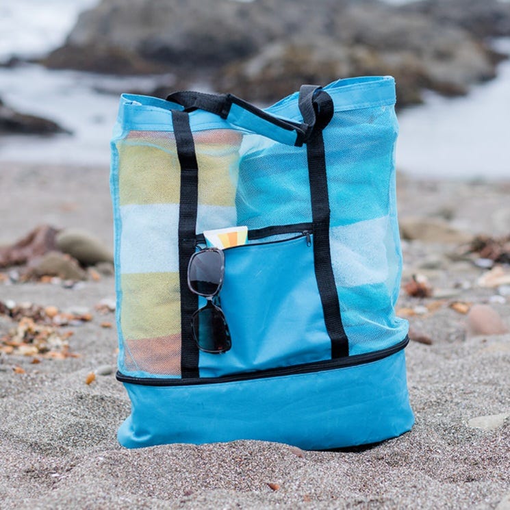 A striped beach bag with sunglasses and sunscreen is on the sand against a rocky beach backdrop.