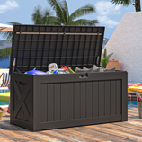Outdoor storage deck box with a hinged lid, shown open, containing various pool toys and cushions, set on a wooden deck by a pool and surrounded by tropical plants.