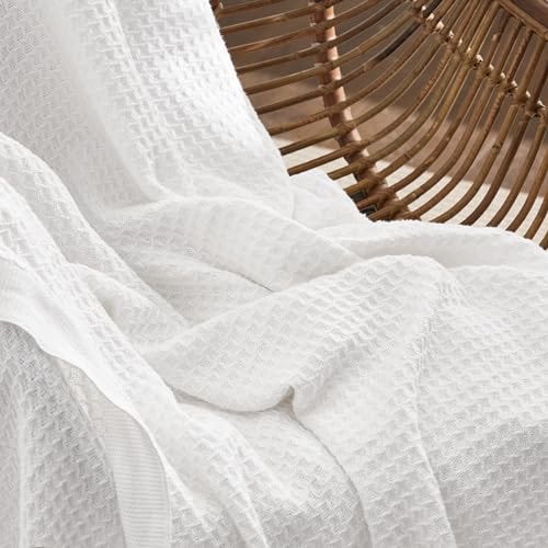 A white waffle weave cotton blanket is casually draped over a wooden hammock, showcasing its textured pattern.