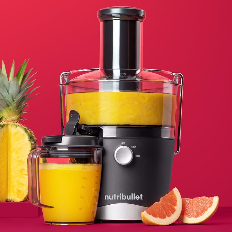 A NutriBullet juicer with a jug of juice, next to a pineapple and grapefruit slices on a red background.